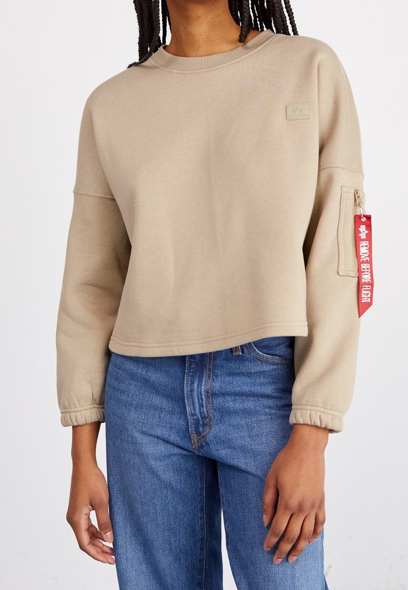 Sweat-shirt court beige à manches longues, col côtelé, et poche zippée avec une étiquette rouge "Remove Before Flight".