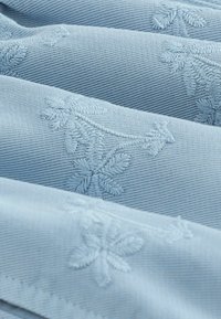 Light blue fabric with a textured weave and subtle floral embroidery. The material appears smooth with a soft sheen, layered in folds.