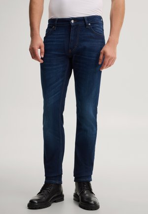 Uomo che indossa jeans slim fit blu scuro, stivali neri con lacci e camicia bianca infilata, in piedi su uno sfondo chiaro e semplice.