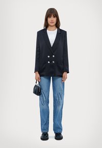 Blazer doppiopetto marina, con rever classici e tasche frontali; abbinato a jeans azzurri chiari con loghi discreti e scarpe con plateau nere.
