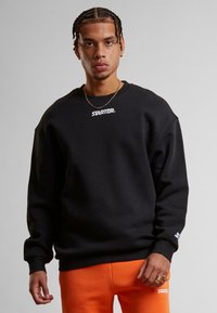 Sweatshirt noir à col rond et épaules tombantes, avec un logo blanc "STARTER" sur la poitrine et des poignets côtelés. Porté avec un pantalon de survêtement orange.