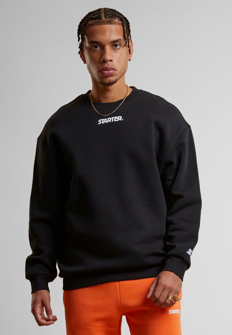 Sweatshirt noir à col rond et épaules tombantes, avec un logo blanc "STARTER" sur la poitrine et des poignets côtelés. Porté avec un pantalon de survêtement orange.