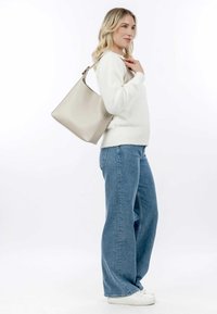 Beige leather handbag with a structured design, large opening, and adjustable strap, worn over a white sweater and blue wide-leg jeans.