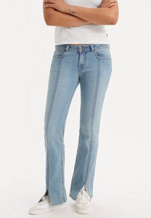 Osoba nosząca jasnoniebieskie dżinsy z detalami szwów z przodu i rozcięciami na kostkach z przodu, połączone z białymi trampkami i białym krótkim topem.