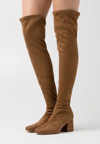 Brown over-the-knee boots made of soft suede, featuring a block heel and a fitted, slouched design. Smooth texture with no visible hardware.