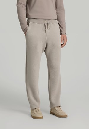 Man wearing beige knit drawstring pants, beige sneakers, and a long-sleeve beige top standing against a light background.