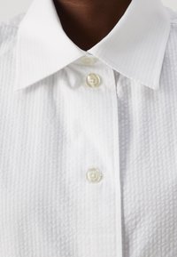Camicia bianca con un tessuto testurizzato. Presenta un colletto classico e tre bottoni perlacei sulla patta frontale.