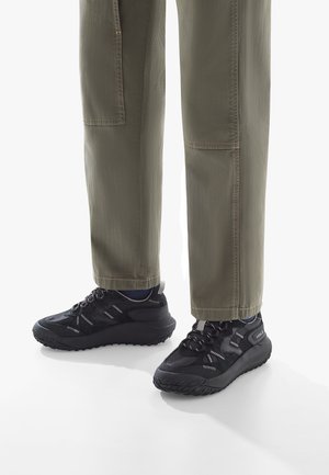 Baskets de sport noires avec un matériau texturé et des semelles robustes, portées avec un pantalon cargo vert olive présentant des coutures visibles et des poches.