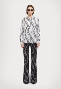 Λευκό πουκάμισο με μακριά μανίκια και μαύρο σχέδιο αλυσίδας, συνδυασμένο με μαύρο παντελόνι καμπάνα που φέρει λευκό κείμενο. Και τα δύο κομμάτια έχουν λεία υφή.