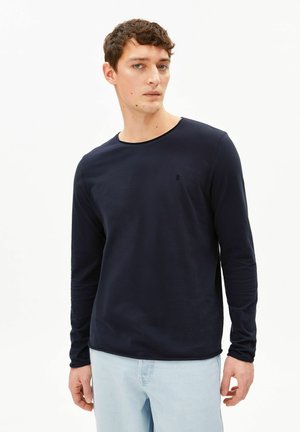 Junger Mann mit kurzen lockigen Haaren, der ein dunkelblaues Langarmshirt und hellblaue Jeans trägt, steht vor einem weißen Hintergrund.