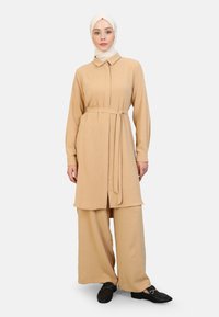 Vestido camisero beige de manga larga con collar y cinturón de lazo, combinado con pantalones de pierna ancha. Textura suave, botones mínimos y zapatos negros.
