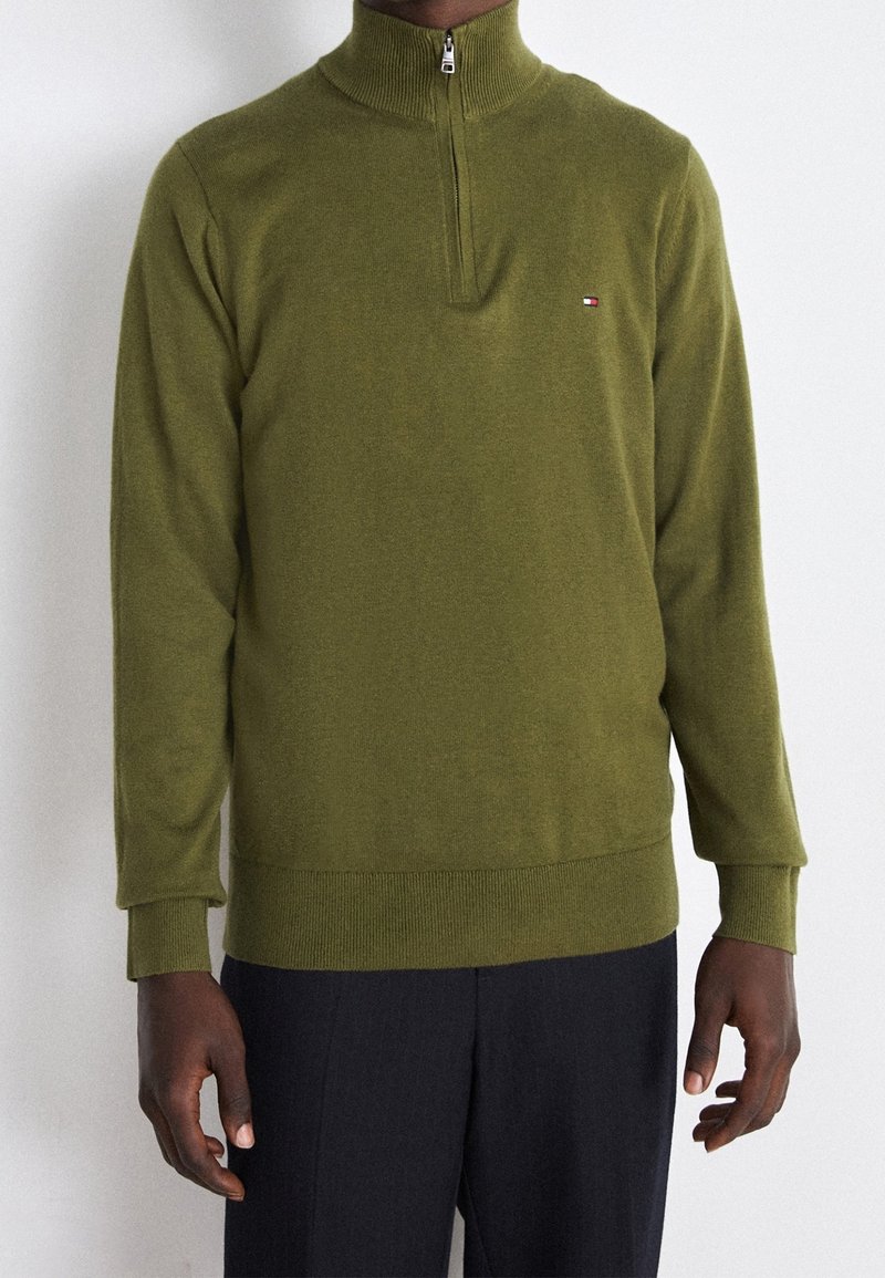Homme portant un pull vert olive à fermeture éclair quart de zip avec petit logo sur la poitrine gauche et un pantalon noir, debout devant un fond clair uni.