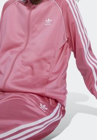 Różowy dres Adidas wykonany z gładkiej tkaniny, z białymi paskami na rękawach i nogawkach, z małym białym logo na piersi i udzie.