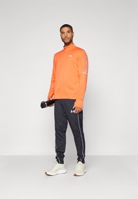 Orange long-sleeve athletic top with half-zip neck; black tapered pants with white accent stripes; beige sneakers; holding a black water bottle.