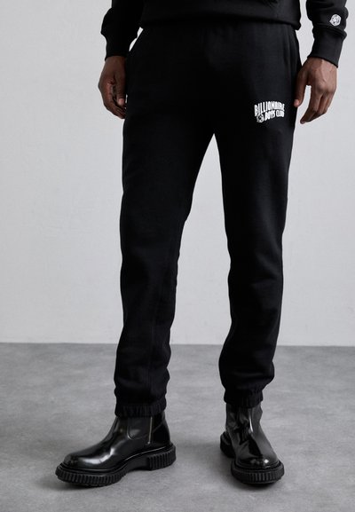 Schwarze Jogginghose aus Baumwolle mit gerippten Bündchen und einem weißen Logo-Druck. Kombiniert mit glänzenden schwarzen Stiefeletten mit einer klobigen Sohle.