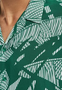 Camicetta a maniche corte di colore verde con un motivo astratto bianco. Include un colletto, chiusura a bottoni e una texture del tessuto morbida.