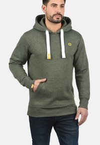 Green hoodie with white drawstrings, kangaroo pocket, and yellow logo. Comfy fabric with ribbed cuffs and hem. Worn by a male model.