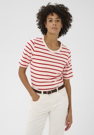 Donna con capelli ricci che indossa una maglietta a maniche corte a righe rosse e bianche, pantaloni bianchi e una cintura marrone, in piedi davanti a uno sfondo uniforme.