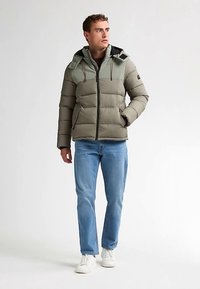 Gewatteerd jack in licht olijfgroen met een donkerder groen paneel op de schouders, ritssluiting en verstelbare capuchon met trekkoord. Gecombineerd met een blauwe spijkerbroek.