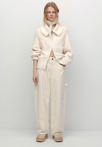 Roomwit gebreid jack met ritssluiting en een brede kraag, gecombineerd met beige wijde broek met hoge taille en plateauzolen. Zachte textuur overal.