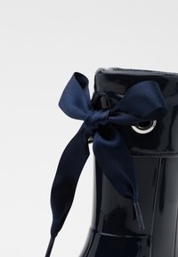 Bottine à tige patent marine avec une finition brillante, ornée d'un grand nœud en satin en accent et d'un détail en œillet métallique près du haut.