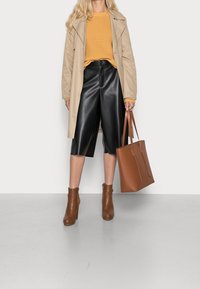 Beżowy trencz na musztardowym swetrze z dzianiny, czarne krótkie spodenki z eko skóry, brązowe botki za kostkę oraz brązowa torba typu tote. Proste tło.