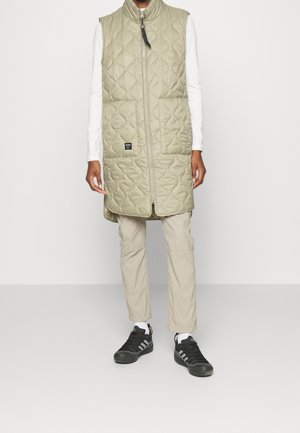 Henkilö, joka on pukeutunut pitkään, tikattuun vaalean oliivinvihreään liiviin valkoisten pitkähihasten päällä, beigenvärisiin housuihin ja mustiin lenkkareihin valkoisten sukkien kera, seisoo paikoillaan.