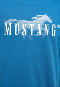 Tissu bleu avec un graphique de cheval stylisé en blanc et le mot « MUSTANG » en grandes lettres majuscules sur la poitrine.