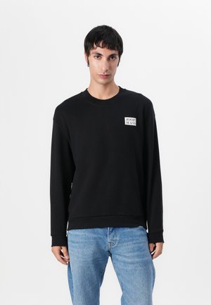 Black sweatshirt with a crew neckline and long sleeves. Features a square logo patch on the chest. Worn with light blue denim jeans.