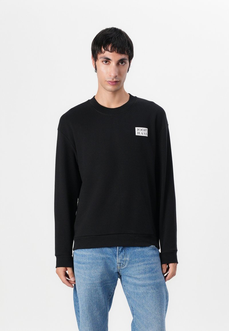 Felpa nera con scollo rotondo e maniche lunghe. Presenta un'etichetta quadrata sul petto. Indossata con jeans in denim blu chiaro.