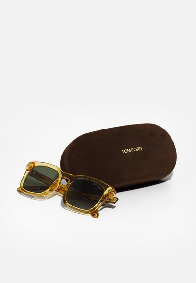 Tom Fordi prillid, millel on läbipaistev kollane raam ja tumerohelised objektiivid, koos pruuni kõvakaanelise karbiga, millel on reljeefne kiri "TOM FORD."