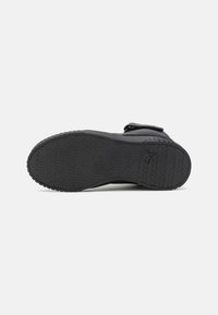 Semelle de chaussure de sport noire avec une surface en caoutchouc texturé, présentant un motif de semelle et un logo en relief sur le côté.
