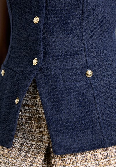 Marinblå texturerad väst med guldknappar och sidofickor, parat med ett brunt rutigt tyg under.