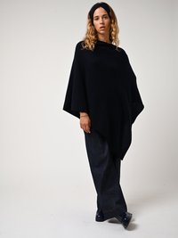 Poncho noir en tricot avec un design drapé, présentant un col haut et des manches larges. Associé à un pantalon large en denim foncé et des chaussures navy.