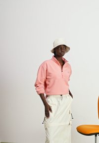 Sudadera de forro polar rosa con cremallera y falda ajustable en un tono blanco roto, combinada con un sombrero tipo bucket en color beis. Tejido suave y corte relajado.