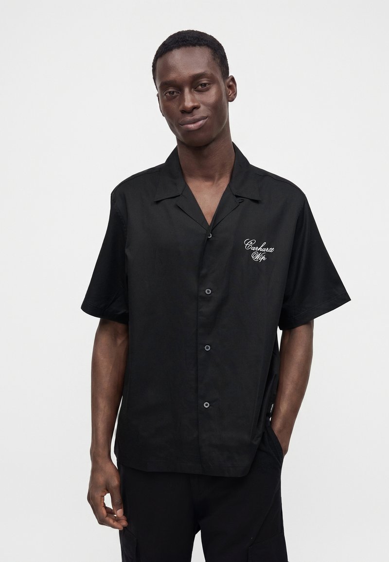Uomo che indossa una camicia nera a maniche corte con testo bianco ricamato sul petto sinistro, in piedi davanti a uno sfondo neutro.