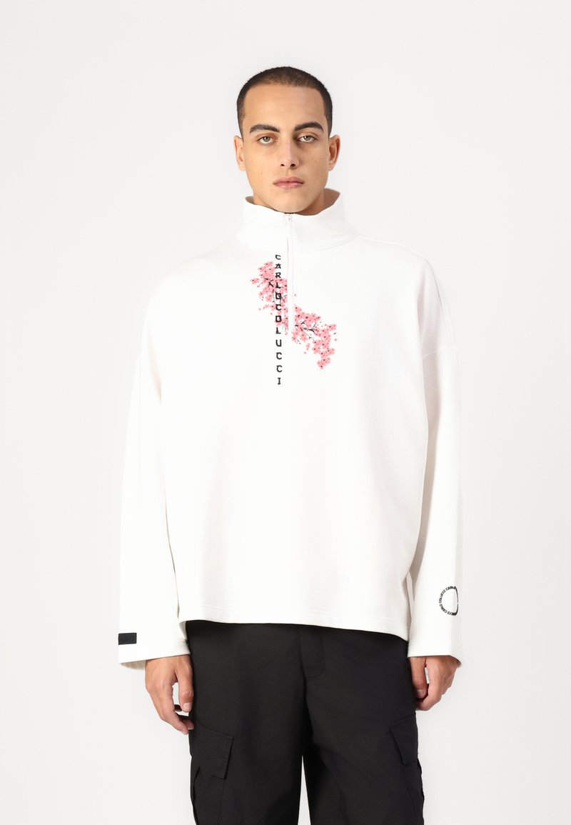 Balta užtrauktuku zip-up megztinis su rožiniu gėlių raštu ir "CARRIEDLUCCI" užrašu. Ilgomis rankovėmis, su juodu akcentu ant rankogalių.