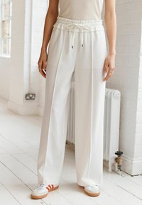 Witte wijde broek met een elastische tailleband en een trekkoord. Gepaard met grijze sneakers met rode strepen.