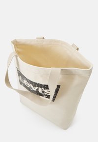 Torba na ramię wykonana z płótna w kolorze kremowym z czarnym graficznym logo. Posiada dwie wytrzymałe rączki oraz otwarte wnętrze, gładką teksturę i prosty design.