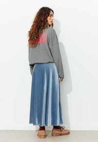 Sweatshirt gris avec texte rose, associé à une jupe en velours légère et fluide de couleur bleu clair et des chaussures épaisses beiges. Vu de côté.
