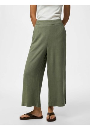 Osoba ubrana w luźne oliwkowozielone skrócone spodnie, biały bezrękawnik i ciemnobrązowe płaskie sandały, stoi na jednolitym tle.