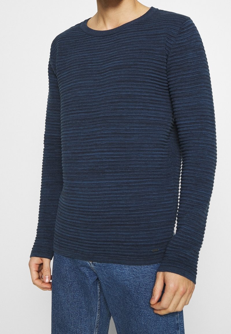 Homme portant un pull côtelé texturé bleu foncé à manches longues et un jean bleu en denim sur un fond uni.