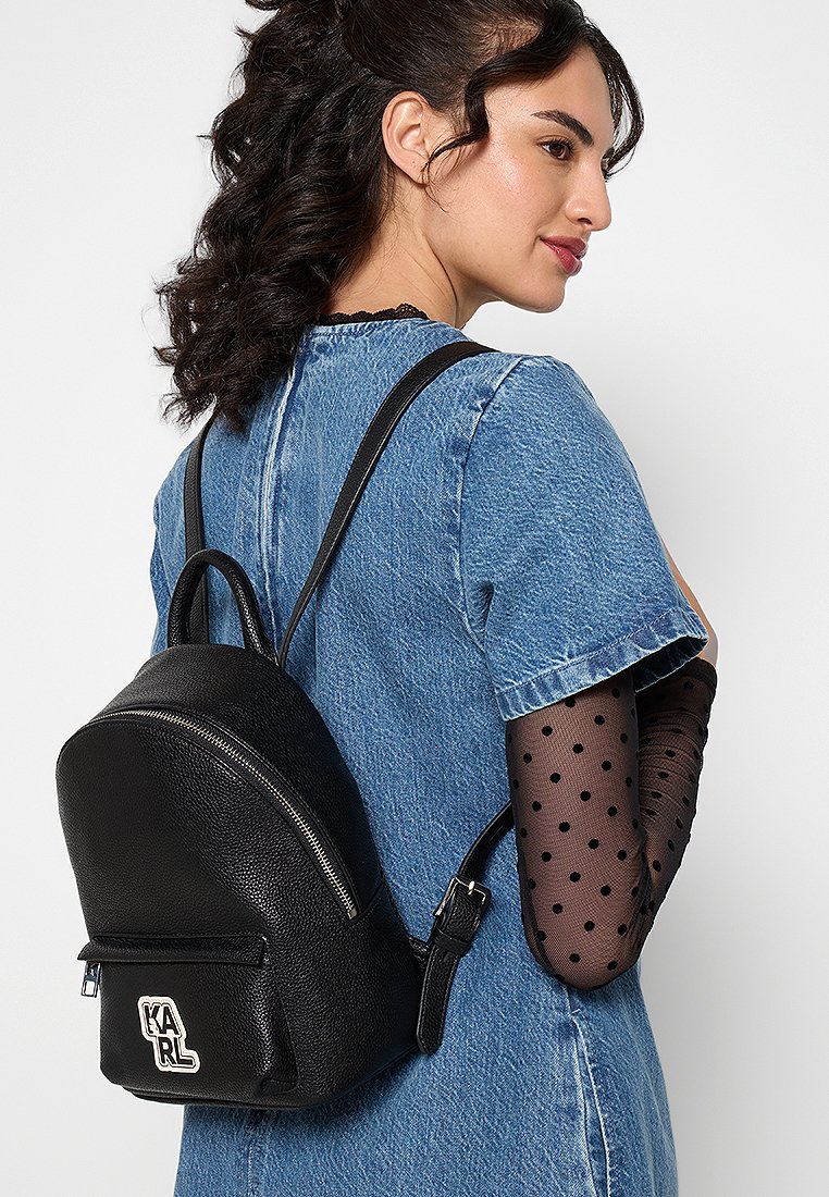 Czarny skórzany plecak z zaokrąglonym dnem, przednią kieszenią na zamek i białym logo. Modelka ma na sobie dżinsową sukienkę z rękawami w kropki.