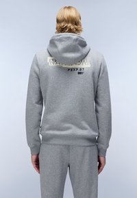 Haut à capuche gris avec une capuche, présentant un logo imprimé crème au dos. Tissu doux avec des poignets et un ourlet côtelés. Pantalon de jogging gris assorti visible.