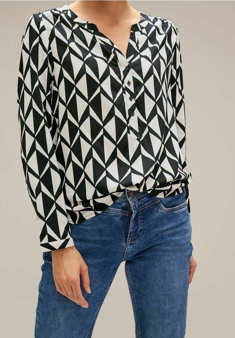 Personne portant un chemisier à manches longues à motifs géométriques noir et blanc, rentré dans un jean en denim bleu, debout devant un fond uni.