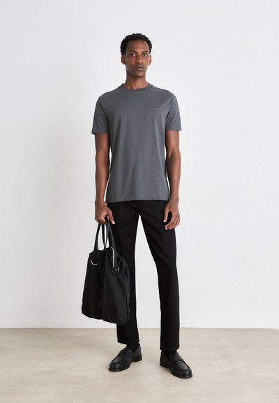 Homme debout contre un mur blanc, portant une chemise grise à manches courtes, un pantalon et des chaussures noirs, tenant un sac fourre-tout noir dans sa main gauche.