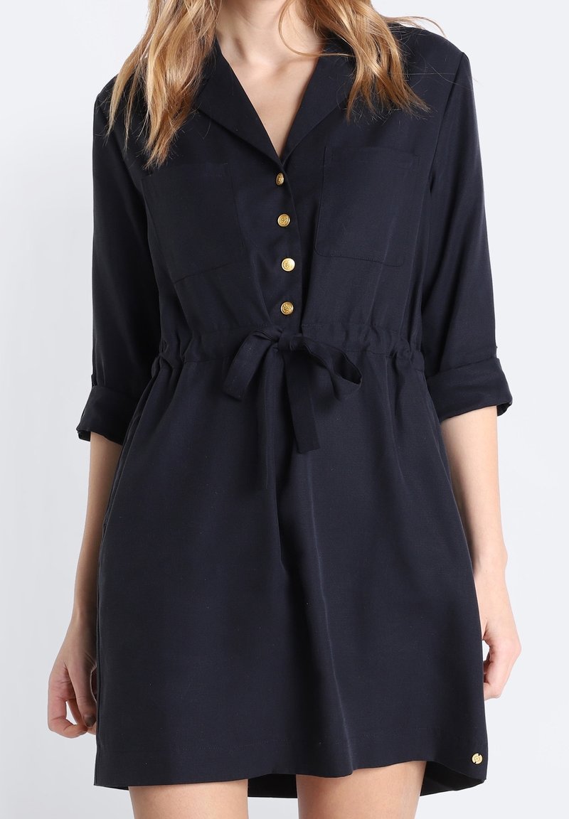 Robe chemise bleu marine avec col, fermeture à boutons sur le devant, deux poches poitrine et une taille nouée. Présente un léger évasement à l'ourlet et des accents dorés.