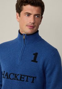 Suéter azul con cremallera tipo cuello de nubosidad. Presenta un número "1" negro y el logotipo de "HACKETT" en negro. Cuello de canalé y corte estándar.