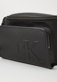 Zwarte leren tas met een ritssluiting bovenaan, een reliëf "CK" logo aan de voorkant en een zijvak met ritssluiting. Soepele textuur.