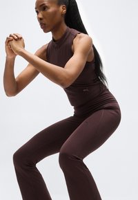 Γυναίκα με σκούρα καφέ αθλητική ένδυση εκτελεί άσκηση κάθισμα με τα χέρια σταυρωμένα μπροστά και έκφραση συγκέντρωσης.